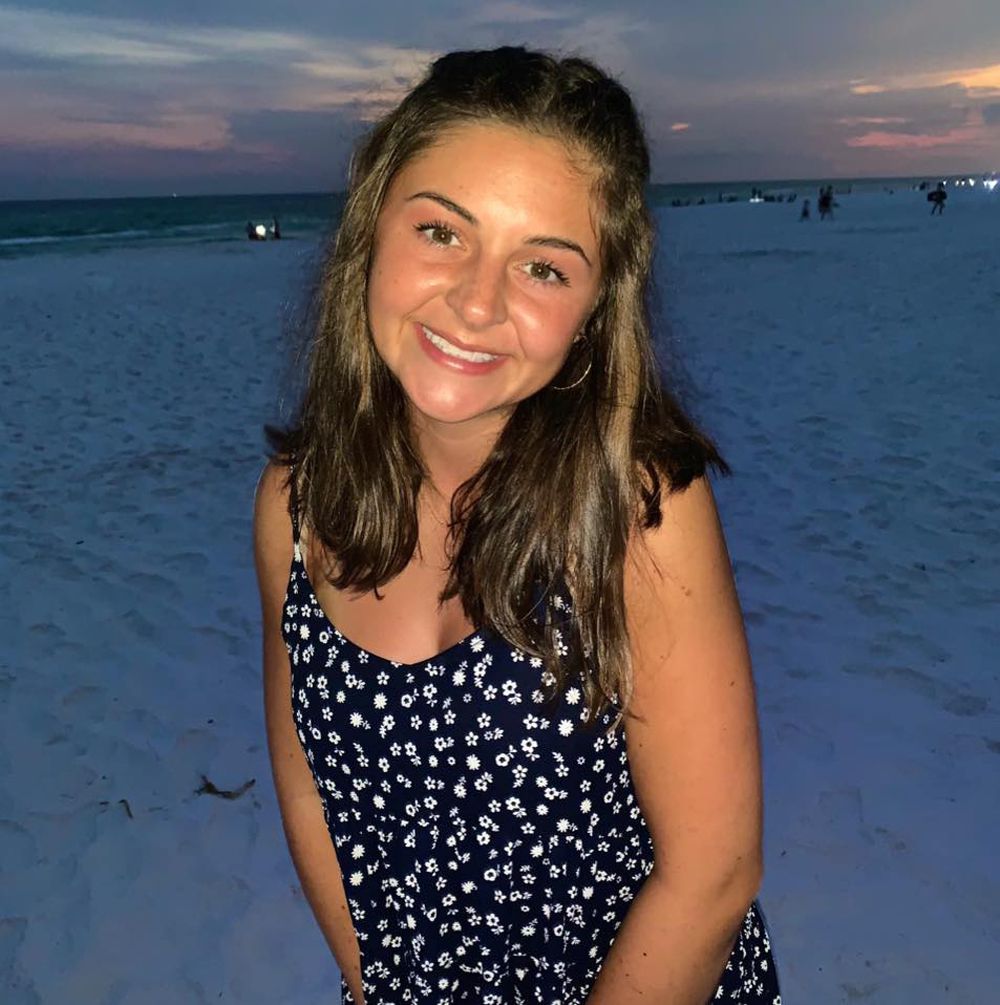 A young woman smiling while standing on a beach during sunset, wearing a floral dress.