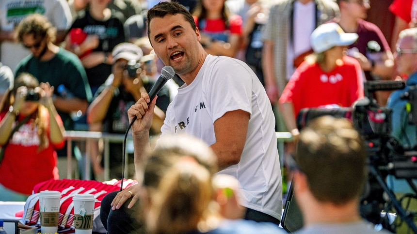 A young man speaking into a microphone during a public event, with a crowd of supporters in the background.
