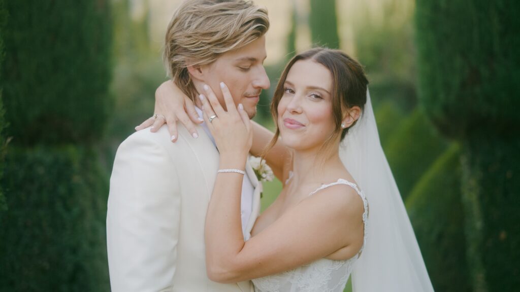 A happy couple embracing in a garden, the woman in a wedding dress and veil, and the man in a white suit, both smiling.